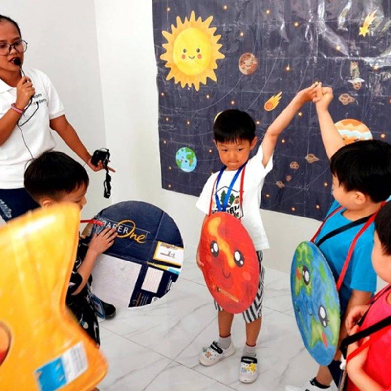 우주 그림 앞에서 행성 모형을 목에 걸고 손을 든 킨더 아이들과 마이크를 든 선생님